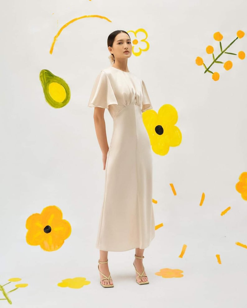 May include: A woman wearing a light beige midi dress with a round neckline and short, flared sleeves. She is standing on a white background with yellow painted flowers and avocado illustrations.