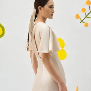 May include: A woman wearing a light beige satin dress with a low back and flutter sleeves. The dress has a tie at the back of the neck. The woman has her hair pulled back in a braid with a light beige ribbon.