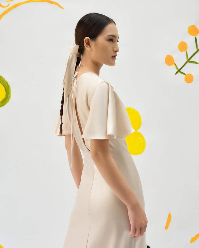 May include: A woman wearing a light beige satin dress with a low back and flutter sleeves. The dress has a tie at the back of the neck. The woman has her hair pulled back in a braid with a light beige ribbon.