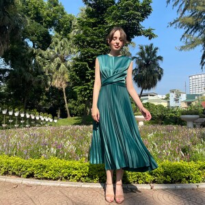 Peut inclure: Une femme portant une robe midi pliss&eacute;e vert sarcelle avec un d&eacute;tail nou&eacute; &agrave; la taille. Elle se tient sur un chemin pav&eacute; de briques dans un jardin.