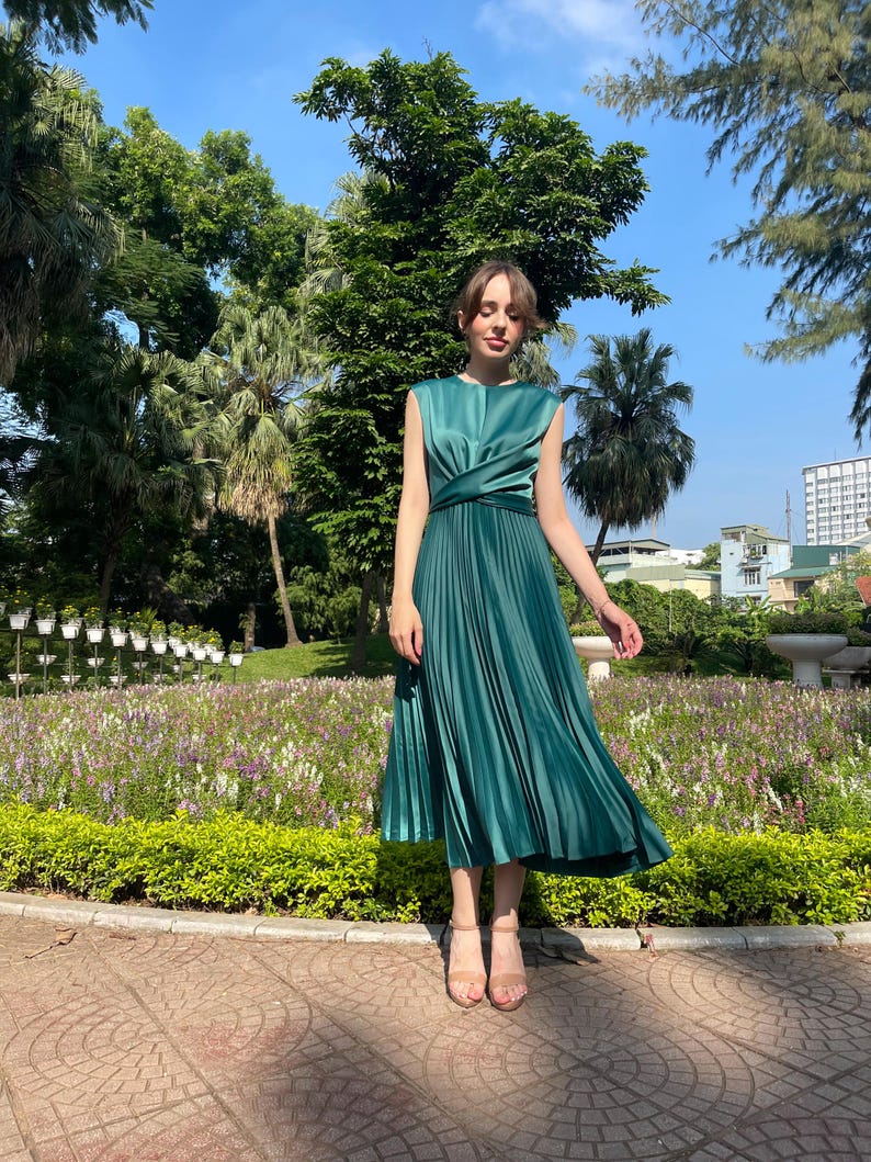 Peut inclure: Une femme portant une robe midi pliss&eacute;e vert sarcelle avec un d&eacute;tail nou&eacute; &agrave; la taille. Elle se tient sur un chemin pav&eacute; de briques dans un jardin.