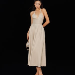 Puede incluir: Un vestido beige con cuello halter y falda fluida. El vestido presenta un corpiño ajustado con pliegues horizontales y un escote pronunciado. La modelo lleva bailarinas beige y un pequeño bolso plateado redondo con correa de cadena.