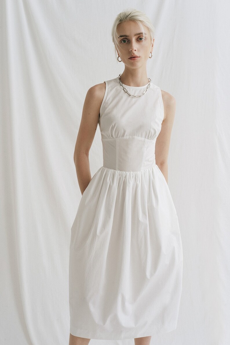 May include: A white sleeveless midi dress with a fitted bodice and a gathered skirt. The dress has a simple, elegant design.