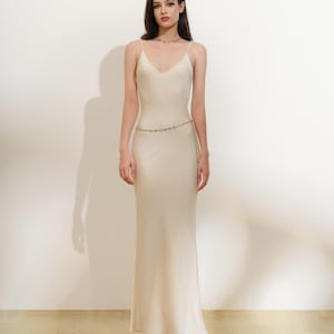 Könnte beinhalten: Ein cremefarbenes Slip-Kleid mit Spaghettiträgern und einem V-Ausschnitt. Das Kleid ist bodenlang und hat einen dezenten Glanz. Ein zarter goldener Kettengürtel betont die Taille. Das Model trägt eine goldene Halskette.