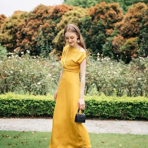 May include: A woman in a yellow maxi dress with a black purse and black sandals stands in a garden setting.