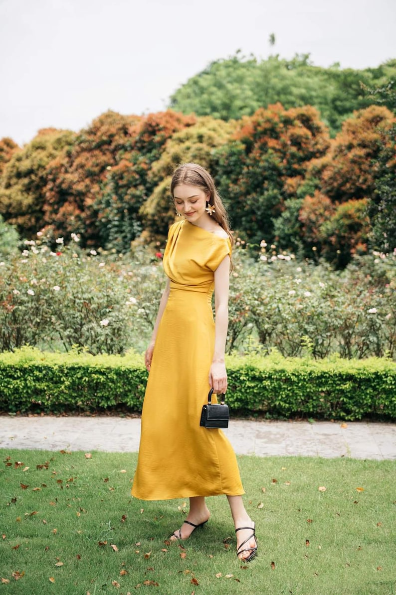 May include: A woman in a yellow maxi dress with a black purse and black sandals stands in a garden setting.