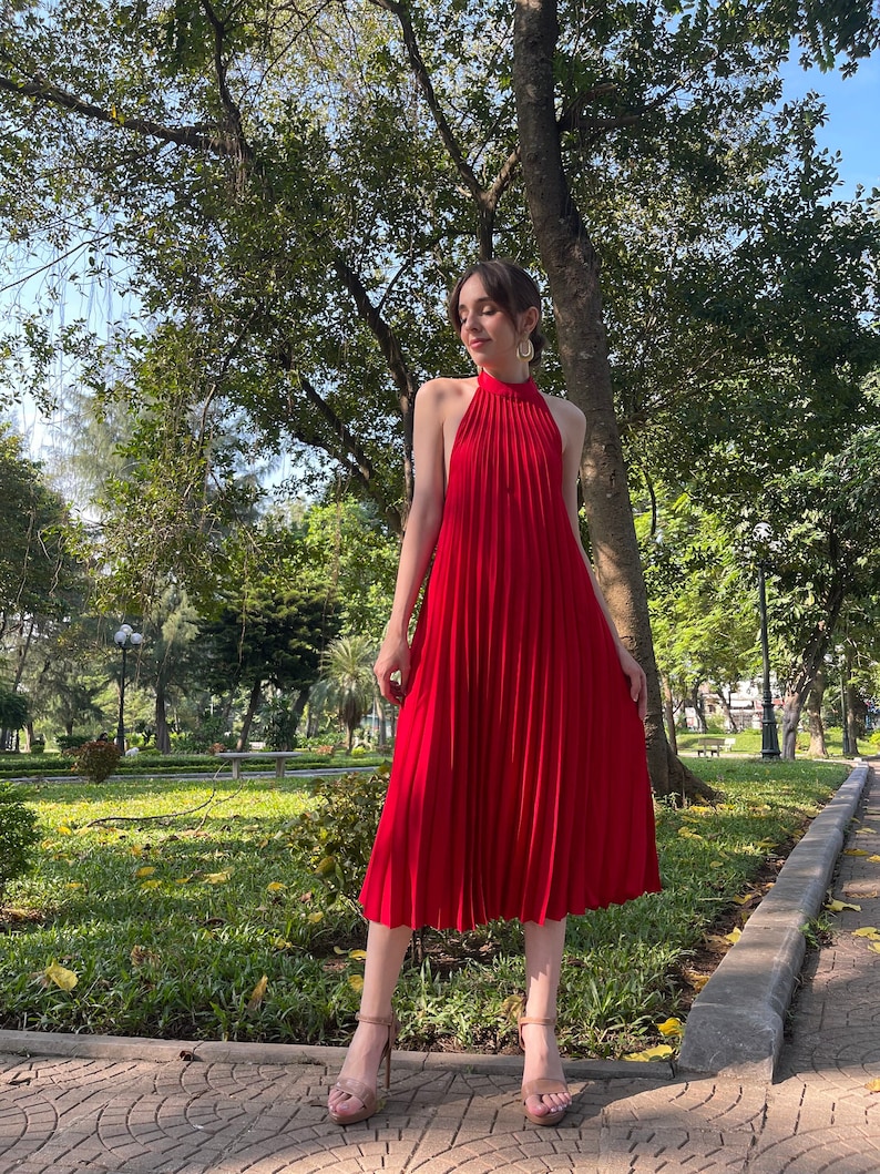 Pode incluir: Uma mulher vestindo um vestido vermelho de al&ccedil;as com pregas. O vestido &eacute; midi e sem mangas. Ela est&aacute; em p&eacute; em um caminho de paralelep&iacute;pedos em um parque.