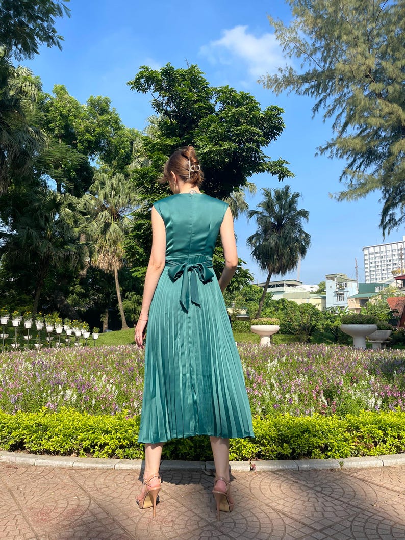 Peut inclure: Une robe midi pliss&eacute;e vert sarcelle avec un n&oelig;ud dans le dos. La robe est sans manches et a un col en V.