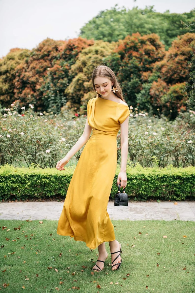 May include: A woman wearing a yellow midi dress with a one-shoulder neckline and a black purse. She is standing in a garden with green grass and flowers.