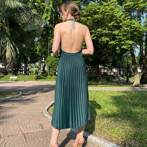 Pode incluir: Um vestido verde escuro de al&ccedil;as com saia plissada. O vestido &eacute; sem mangas e tem costas baixas. A modelo est&aacute; usando o vestido com saltos nude.