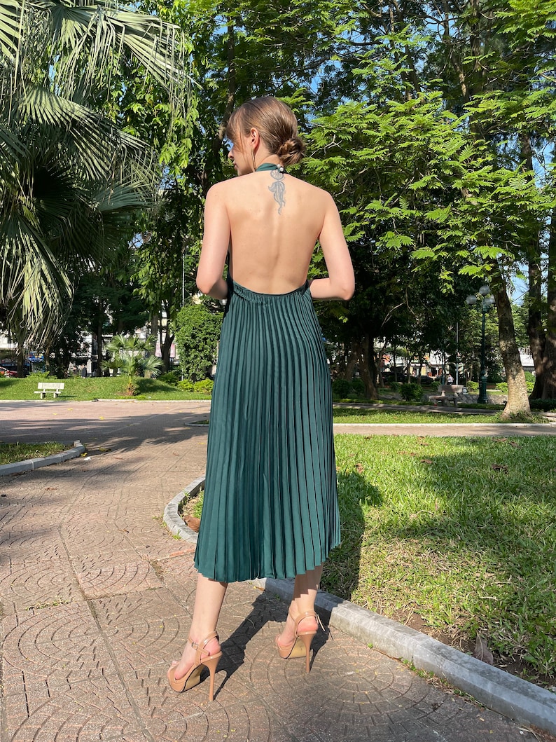 Pode incluir: Um vestido verde escuro de al&ccedil;as com saia plissada. O vestido &eacute; sem mangas e tem costas baixas. A modelo est&aacute; usando o vestido com saltos nude.