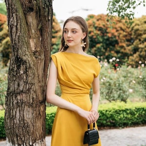May include: A woman in a mustard yellow dress with a black purse stands next to a tree. She is wearing gold flower earrings.