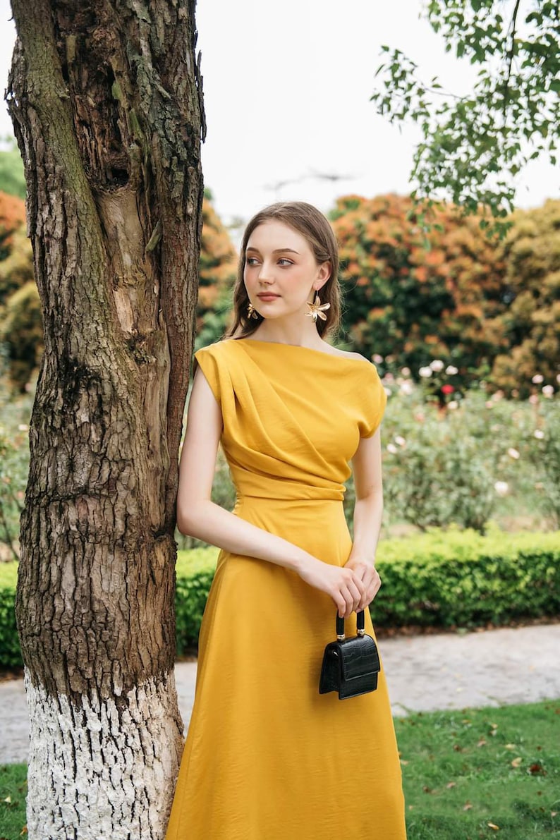 May include: A woman in a mustard yellow dress with a black purse stands next to a tree. She is wearing gold flower earrings.