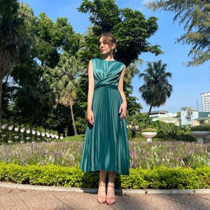Peut inclure: Une robe midi pliss&eacute;e verte avec un corsage sans manches et un d&eacute;tail enroul&eacute; &agrave; la taille. La robe est port&eacute;e par une personne debout dans un jardin.