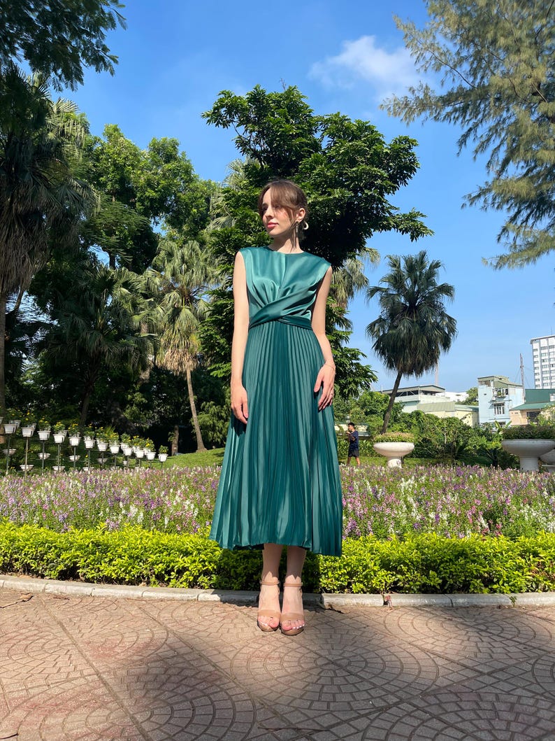 Peut inclure: Une robe midi pliss&eacute;e verte avec un corsage sans manches et un d&eacute;tail enroul&eacute; &agrave; la taille. La robe est port&eacute;e par une personne debout dans un jardin.