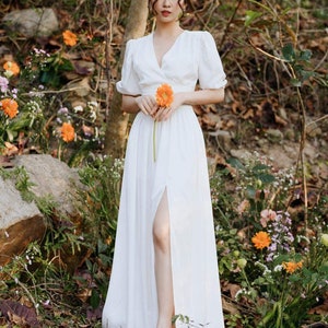 May include: A white maxi dress with a V-neckline, short puff sleeves, and a thigh-high slit. The dress is made of a lightweight fabric and has a flowy silhouette. The model is wearing a flower crown and holding a single orange flower.