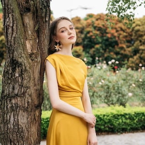 May include: A woman wearing a mustard yellow dress with a black purse and gold star-shaped earrings. She is standing next to a tree in a garden setting.