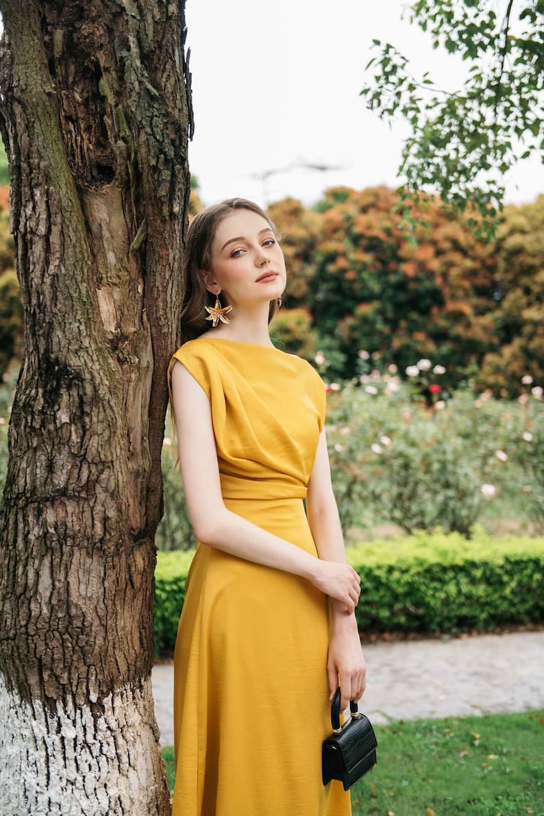May include: A woman wearing a mustard yellow dress with a black purse and gold star-shaped earrings. She is standing next to a tree in a garden setting.