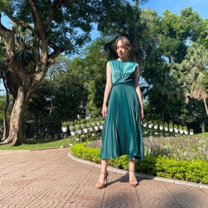 Peut inclure: Une femme portant une robe pliss&eacute;e vert sarcelle avec un corsage torsad&eacute;. Elle se tient dans un parc avec des arbres verts et des fleurs en arri&egrave;re-plan.