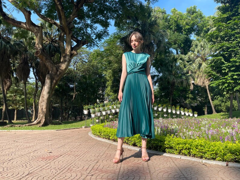 Peut inclure: Une femme portant une robe pliss&eacute;e vert sarcelle avec un corsage torsad&eacute;. Elle se tient dans un parc avec des arbres verts et des fleurs en arri&egrave;re-plan.