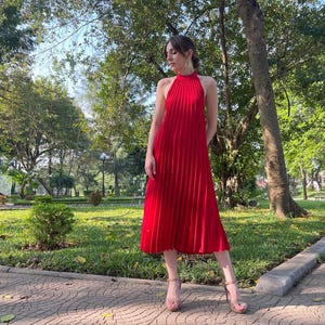 May include: A woman wearing a red pleated halter dress and gold heeled sandals. She is standing on a paved walkway in a park setting.