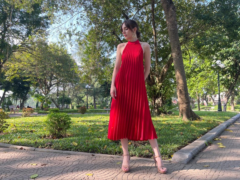 May include: A woman wearing a red pleated halter dress and gold heeled sandals. She is standing on a paved walkway in a park setting.