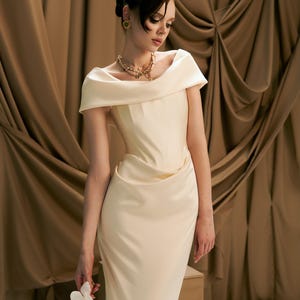 May include: An off-white, floor-length gown with an off-the-shoulder neckline. The dress features a draped bodice and a fitted skirt. A gold necklace and earrings complement the dress. A white flower is held in the hand.