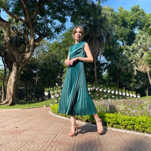Peut inclure: Une femme portant une robe pliss&eacute;e verte et des talons hauts se tient dans un parc. La robe est sans manches et a un corsage ajust&eacute;. La femme sourit et regarde la cam&eacute;ra.