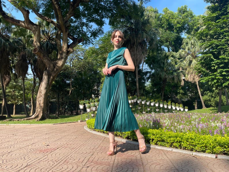 Peut inclure: Une femme portant une robe pliss&eacute;e verte et des talons hauts se tient dans un parc. La robe est sans manches et a un corsage ajust&eacute;. La femme sourit et regarde la cam&eacute;ra.