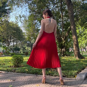 May include: A woman wearing a red pleated dress with a halter neckline and a low back. She is standing in a park with trees and grass in the background.