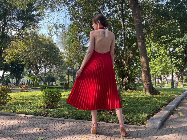 May include: A woman wearing a red pleated dress with a halter neckline and a low back. She is standing in a park with trees and grass in the background.