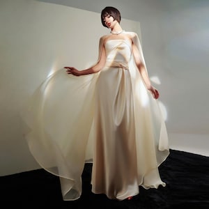 May include: A cream-colored, strapless gown with a flowing cape. The dress features a fitted bodice and a long, elegant skirt. The cape is made of a sheer, lightweight fabric that drapes gracefully. The overall look is sophisticated and ethereal.