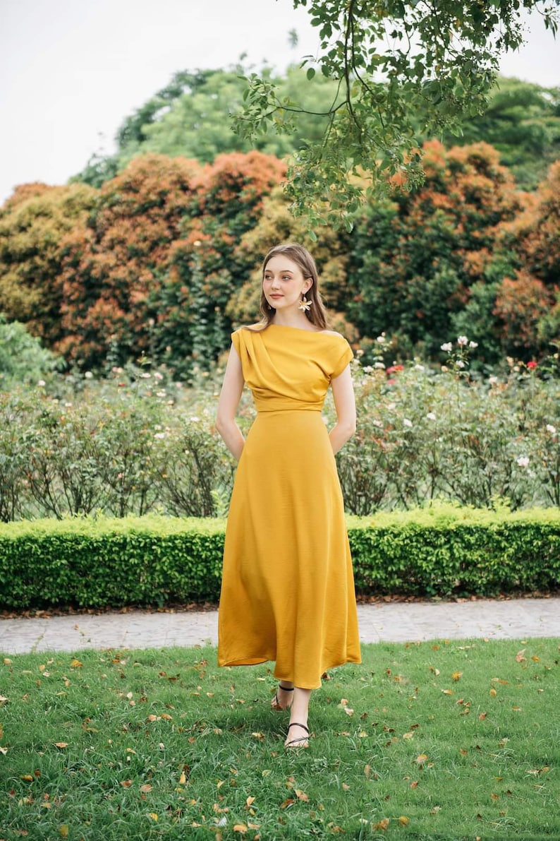 May include: A woman wearing a mustard yellow maxi dress with a draped neckline and short sleeves. She is standing in a garden with green grass and bushes.