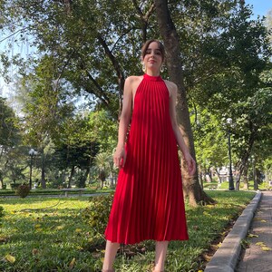 Pode incluir: Um vestido midi vermelho com decote halter e saia plissada. O vestido &eacute; usado por uma pessoa em p&eacute; em um caminho de paralelep&iacute;pedos em um parque.
