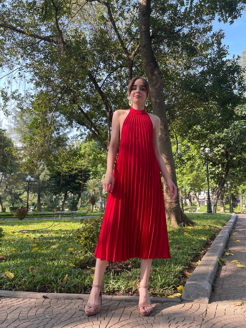 Pode incluir: Um vestido midi vermelho com decote halter e saia plissada. O vestido &eacute; usado por uma pessoa em p&eacute; em um caminho de paralelep&iacute;pedos em um parque.