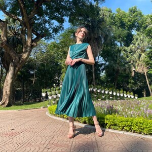 Peut inclure: Une femme portant une robe midi pliss&eacute;e verte sans manches. Elle se tient sur un chemin pav&eacute; de briques dans un parc.
