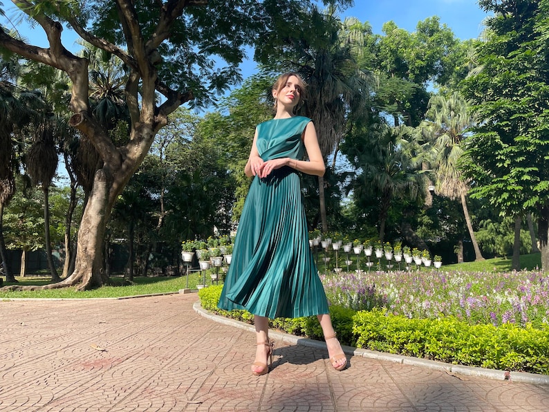 Peut inclure: Une femme portant une robe midi pliss&eacute;e verte sans manches. Elle se tient sur un chemin pav&eacute; de briques dans un parc.