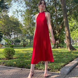 May include: A woman wearing a red halter dress with pleats, standing on a brick walkway in a park. She is wearing gold hoop earrings and nude heels.
