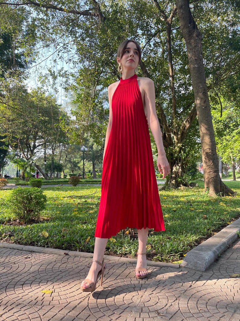 May include: A woman wearing a red halter dress with pleats, standing on a brick walkway in a park. She is wearing gold hoop earrings and nude heels.