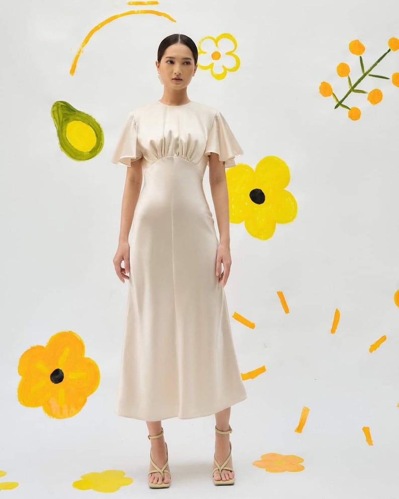 May include: A woman wearing a white satin dress with a round neckline and short, flared sleeves. The dress has a fitted bodice and a flowing skirt. She is wearing beige heeled sandals.