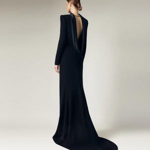 Könnte beinhalten: Ein schwarzes, langärmliges Kleid mit einem Cowl-Ausschnitt und einem tiefen Rücken. Das Kleid hat ein figurbetontes Oberteil und einen fließenden Rock.