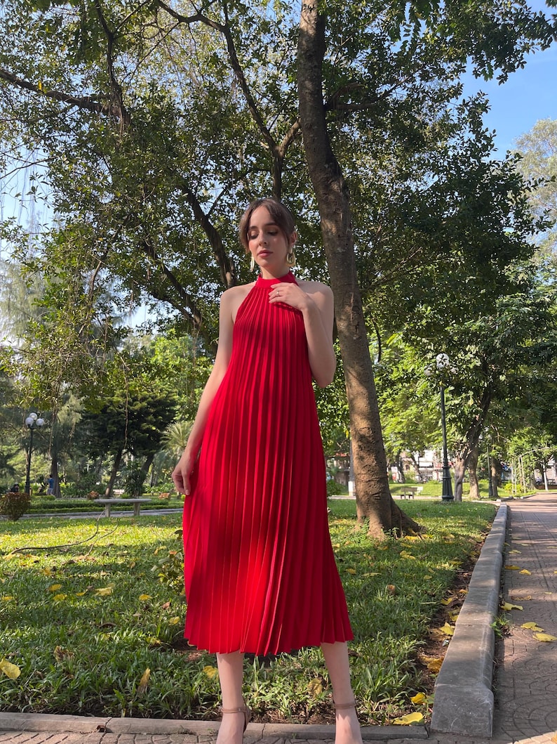 May include: A red pleated midi dress with a halter neckline. The dress is worn by a person standing in a park setting.