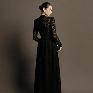 May include: A long, black lace dress with long sleeves and a high neckline. The dress has a fitted bodice and a flowing skirt. The person is holding a small black clutch. The background is a solid, neutral color.