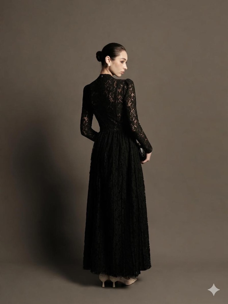 May include: A long, black lace dress with long sleeves and a high neckline. The dress has a fitted bodice and a flowing skirt. The person is holding a small black clutch. The background is a solid, neutral color.
