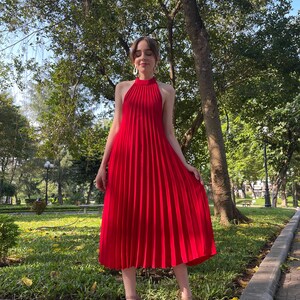 May include: A red midi dress with a halter neckline and pleated skirt. The dress is worn by a person standing on a brick pathway in a park.