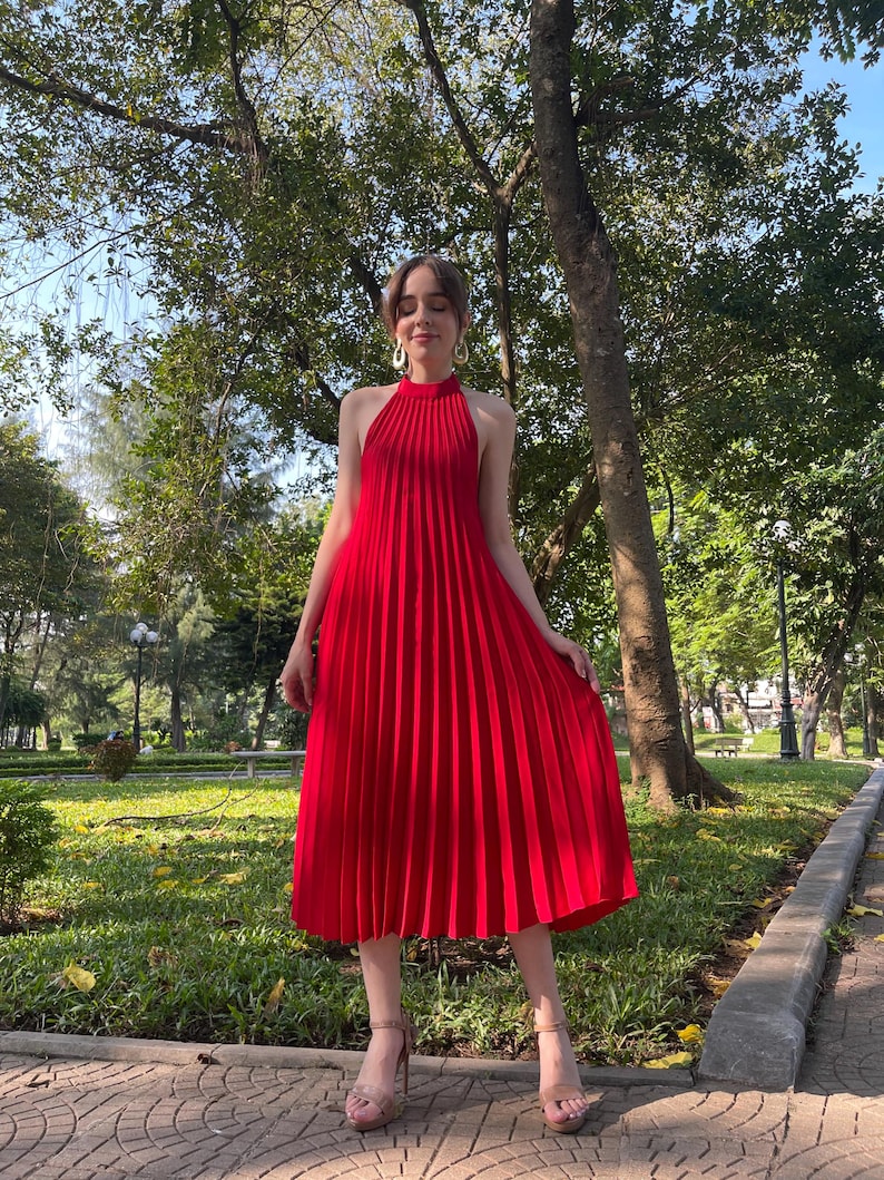 May include: A red midi dress with a halter neckline and pleated skirt. The dress is worn by a person standing on a brick pathway in a park.