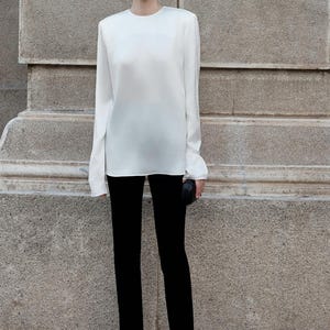 May include: A white long-sleeved blouse with a round neckline and black pants. The pants have a slit at the bottom of each leg.