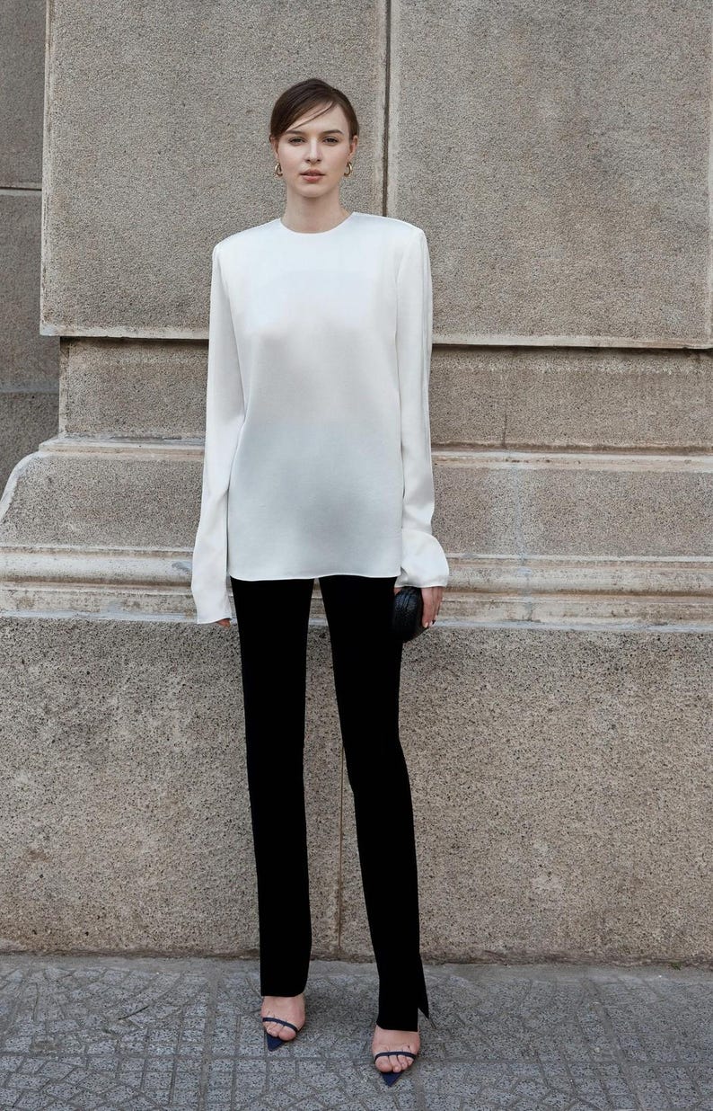 May include: A white long-sleeved blouse with a round neckline and black pants. The pants have a slit at the bottom of each leg.