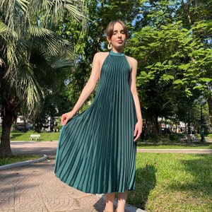 Pode incluir: Uma mulher vestindo um vestido midi plissado verde escuro com decote halter. Ela est&aacute; em p&eacute; em uma cal&ccedil;ada pavimentada em um parque.