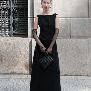 May include: A black sleeveless maxi dress with a boat neckline. The outfit includes sheer black gloves, a woven clutch, and gold hoop earrings. The model is standing in front of a stone wall.